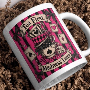 K&ouml;nnte beinhalten: Ein wei&szlig;er Keramikbecher mit pink-schwarzem Streifenhintergrund und einem Stapel aus drei Teetassen, die mit Herz-, Karo- und Pik-Symbolen verziert sind. Der Text lautet "Tea First, MADNESS Later", erg&auml;nzt durch Spielkarten, W&uuml;rfel und Schleifen.