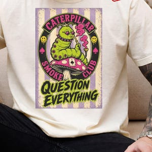 K&ouml;nnte beinhalten: Ein cremefarbenes T-Shirt mit einer Grafik einer gr&uuml;nen Raupe, die eine rosa Brille tr&auml;gt und an einer Pfeife raucht, w&auml;hrend sie auf einem rosa Pilz sitzt. Das Design enth&auml;lt den Text CATERPILLAR SMOKE CLUB und QUESTION EVERYTHING.