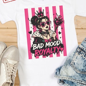 K&ouml;nnte beinhalten: Ein wei&szlig;es T-Shirt mit einer Grafik einer Person, die eine Krone, herzf&ouml;rmige Sonnenbrillen und eine Jacke mit Pelzkragen tr&auml;gt und ein herzf&ouml;rmiges Zepter h&auml;lt. Das Design zeigt rosa vertikale Streifen und den Text BAD MOOD ROYALTY in Schwarz und Pink.