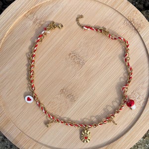 Peut inclure: Un collier doré avec des accents tissés rouges et blancs. Le collier comporte divers breloques, dont un cœur, un champignon, une étoile et un pendentif rectangulaire avec un motif de cœur. Le collier est présenté sur un plateau en bois.