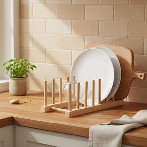 May include: A wooden plate rack holds two white plates and a clear glass. A wooden cutting board rests behind the plates. The rack sits on a wooden countertop, with a small potted plant and a beige tile backsplash in the background.
