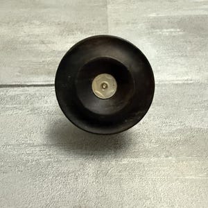 May include: A dark brown bottle stopper with a circular design. The stopper has a flat, wide base and a smaller, raised center with a light-colored, circular inlay. The object is set against a gray background.