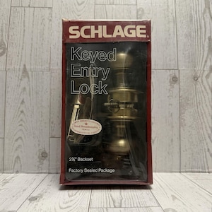 May include: A factory-sealed Schlage keyed entry lock in its original packaging. The box is red and black, with the brand name at the top. The lock is brass-colored, and the package includes a 2 3/8" backset.