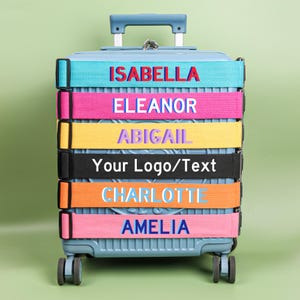 Może przedstawiać: Jasnoniebieska walizka na kółkach z wysuwanym uchwytem. Walizka posiada kolorowe paski z imionami takimi jak "ISABELLA" i "ELEANOR" w różnych kolorach. Tekst "Your Logo/Text" znajduje się na czarnym pasku. Walizka ma cztery koła.