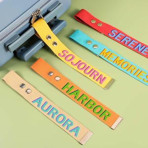 May include: A collection of colorful luggage tags with text, including "AURORA", "HARBOR", "SOJOURN", "MEMORIES", and "SERENE". The tags are in various colors: beige, orange, yellow, teal, and red. Each tag has metal hardware and is next to a blue suitcase.