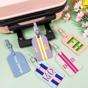 May include: Several personalised luggage tags in shades of blue, lilac, and pink. Each tag displays initials or the text "Your Design". The leather tags have buckle fastenings. The tags are shown next to a pink suitcase.