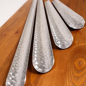 May include: Four silver-colored shoe horns with a textured, hammered metal finish are arranged on a wooden surface. The shoe horns are long and slender, with a rounded end for easy shoe wear.