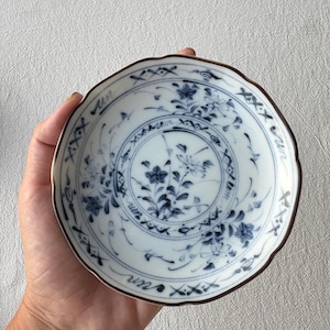 Puede incluir: Plato pequeño y redondo de cerámica blanca con borde festoneado y borde marrón. Presenta un diseño floral azul con una flor central y un motivo de mariposa. La palabra "un" está escrita alrededor del borde.