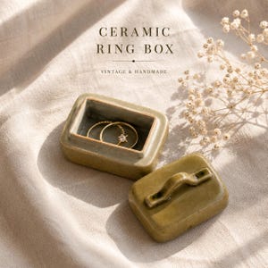 May include: A rectangular olive green ceramic ring box with a matching lid featuring a small handle. The box is open, revealing two gold rings inside. The text CERAMIC RING BOX VINTAGE & HANDMADE is printed in dark brown above the container.