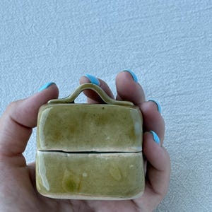May include: A small, olive-green ceramic box with a handle, held in a hand. The box is rectangular with a slightly glossy finish. The handle is arched and integrated into the top. The box is likely used for storing small items.