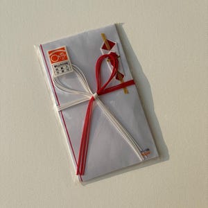 May include: White envelope with red and white decorative string, sealed in clear plastic. The envelope has Japanese text and a red and gold seal. A small red and gold paper decoration is attached.