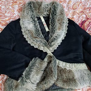 K&ouml;nnte beinhalten: Schwarze Jacke mit Kunstfellkragen und -besatz. Der Kragen und die Manschetten sind mit grauem Kunstfell und Spitze verziert. Die Jacke hat einen einzelnen Knopfverschluss und ein Etikett mit der Aufschrift "E <3 S".