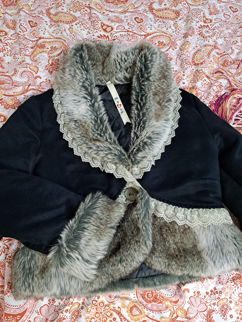 K&ouml;nnte beinhalten: Schwarze Jacke mit Kunstfellkragen und -besatz. Der Kragen und die Manschetten sind mit grauem Kunstfell und Spitze verziert. Die Jacke hat einen einzelnen Knopfverschluss und ein Etikett mit der Aufschrift "E <3 S".
