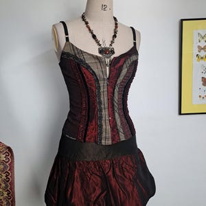 Puede incluir: Vestido estilo corsé burdeos y negro sobre maniquí, con falda en capas. El corpiño combina tejidos de cuadros y encaje, complementado con un collar de cuentas. La falda tiene un diseño texturizado en forma de burbuja. Un artículo de moda único.
