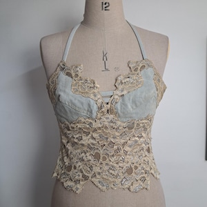 May include: A halter top featuring light blue denim fabric panels combined with intricate beige lace overlays. The garment has a sweetheart neckline, thin straps, and a scalloped hemline. The lace extends over the denim cups and bodice.