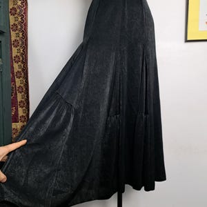 Op de afbeelding: Een zwarte, vloeiende rok met een hoge taille en een getextureerde, licht verweerde uitstraling. De rok heeft een uitlopend silhouet en wordt op een mannequin getoond. De stof lijkt een subtiele glans te hebben.