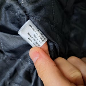K&ouml;nnte beinhalten: Nahaufnahme eines wei&szlig;en Etiketts mit Pflegehinweisen. Das Etikett befindet sich auf einem schwarzen, gesteppten Stoff. Das Etikett lautet: HANDW&Auml;SCHE, NICHT BLEICHEN, B&Uuml;GELN BEI NIEDRIGER TEMPERATUR, NICHT IM TROCKNER TROCKNEN, REINIGEN.