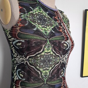 May include: A sleeveless top featuring a repeating butterfly wing pattern in shades of dark brown, green, and blue. The garment has a round neckline with a light purple trim and is displayed on a beige dress form against a plain white wall.