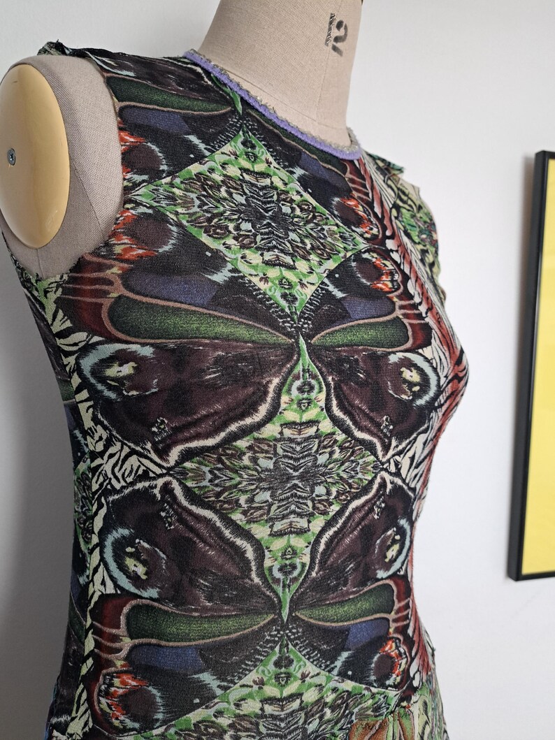 May include: A sleeveless top featuring a repeating butterfly wing pattern in shades of dark brown, green, and blue. The garment has a round neckline with a light purple trim and is displayed on a beige dress form against a plain white wall.