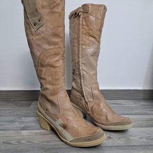 May include: A pair of tan leather knee-high boots. The boots have a distressed appearance with a side zip and a block heel. The left boot has the text "300 Avenue Street YCR" printed on the side.