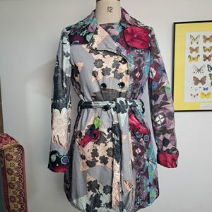 May include: A double-breasted trench coat featuring a patchwork design with various floral and geometric patterns in shades of grey, pink, purple, and black. The coat includes a matching tie belt, a notched collar, and long sleeves with cuffs.