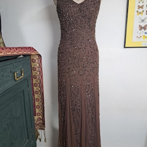 May include: A floor-length brown sleeveless gown featuring a V-neckline and intricate dark beadwork in a swirling vine pattern. The dress is constructed from a sheer overlay fabric with a fitted bodice that transitions into a flowing skirt.
