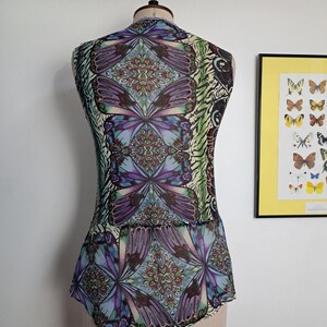 May include: A sleeveless top on a mannequin featuring a vibrant, repeating butterfly wing pattern in shades of purple, blue, and green. The design includes intricate, symmetrical mandala-like motifs integrated within the butterfly wing graphics.