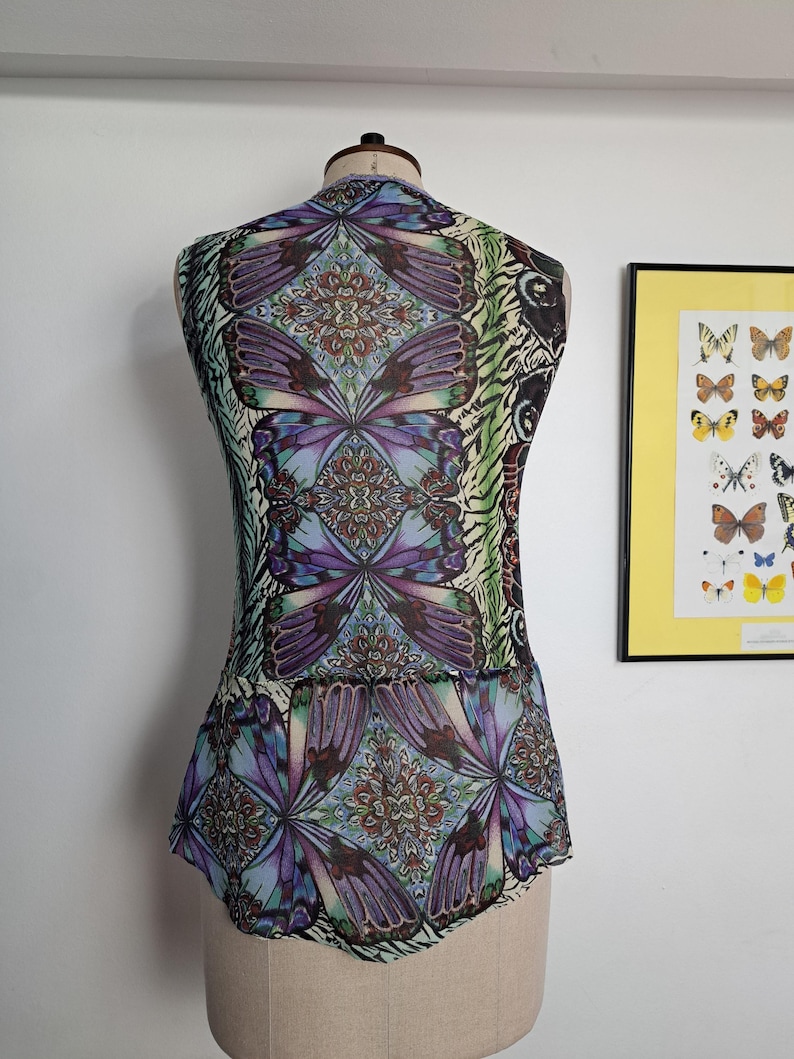 May include: A sleeveless top on a mannequin featuring a vibrant, repeating butterfly wing pattern in shades of purple, blue, and green. The design includes intricate, symmetrical mandala-like motifs integrated within the butterfly wing graphics.