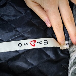 K&ouml;nnte beinhalten: Nahaufnahme eines schwarzen, gesteppten Stoffes mit einem wei&szlig;en Etikett. Das Etikett tr&auml;gt den Text "e ♡ yM" in schwarzen Buchstaben mit einem roten Herzsymbol. Der Stoff ist Teil eines Kleidungsst&uuml;cks.