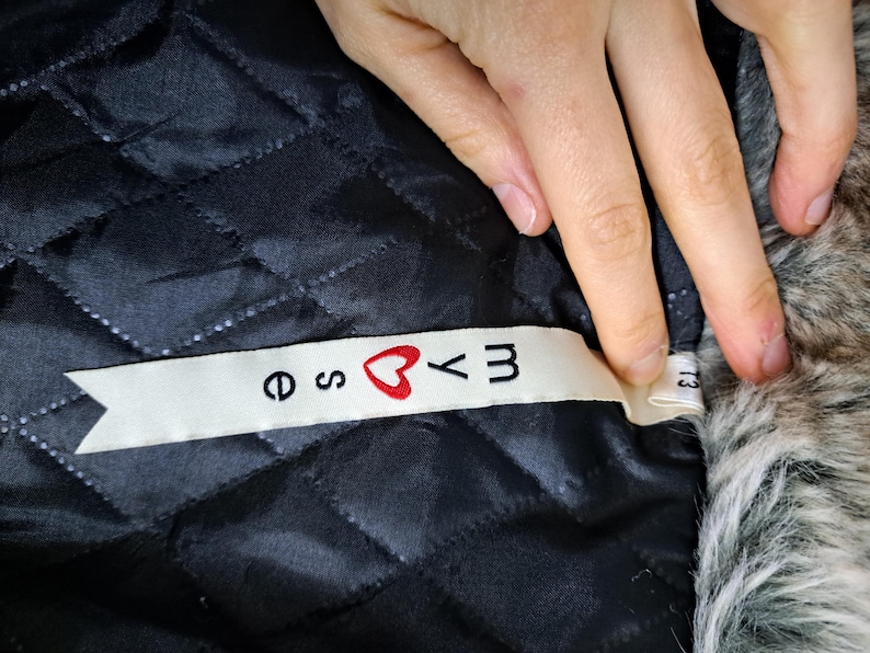 K&ouml;nnte beinhalten: Nahaufnahme eines schwarzen, gesteppten Stoffes mit einem wei&szlig;en Etikett. Das Etikett tr&auml;gt den Text "e ♡ yM" in schwarzen Buchstaben mit einem roten Herzsymbol. Der Stoff ist Teil eines Kleidungsst&uuml;cks.