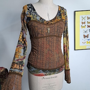 May include: A long-sleeved top with a brown lace bodice and patterned sleeves. The top features a scoop neckline and a decorative clasp. The sleeves have a colorful, vintage-style print with brown lace cuffs. The garment is displayed on a mannequin.