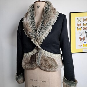 K&ouml;nnte beinhalten: Schwarze Jacke mit Kunstfellbesatz und Kragen. Die Jacke hat eine dekorative Spitzenborte entlang der Kanten des Fells und einen einzelnen Knopfverschluss. Die Manschetten sind ebenfalls mit Kunstfell besetzt. Die Jacke wird auf einer Schneiderpuppe pr&auml;sentiert.
