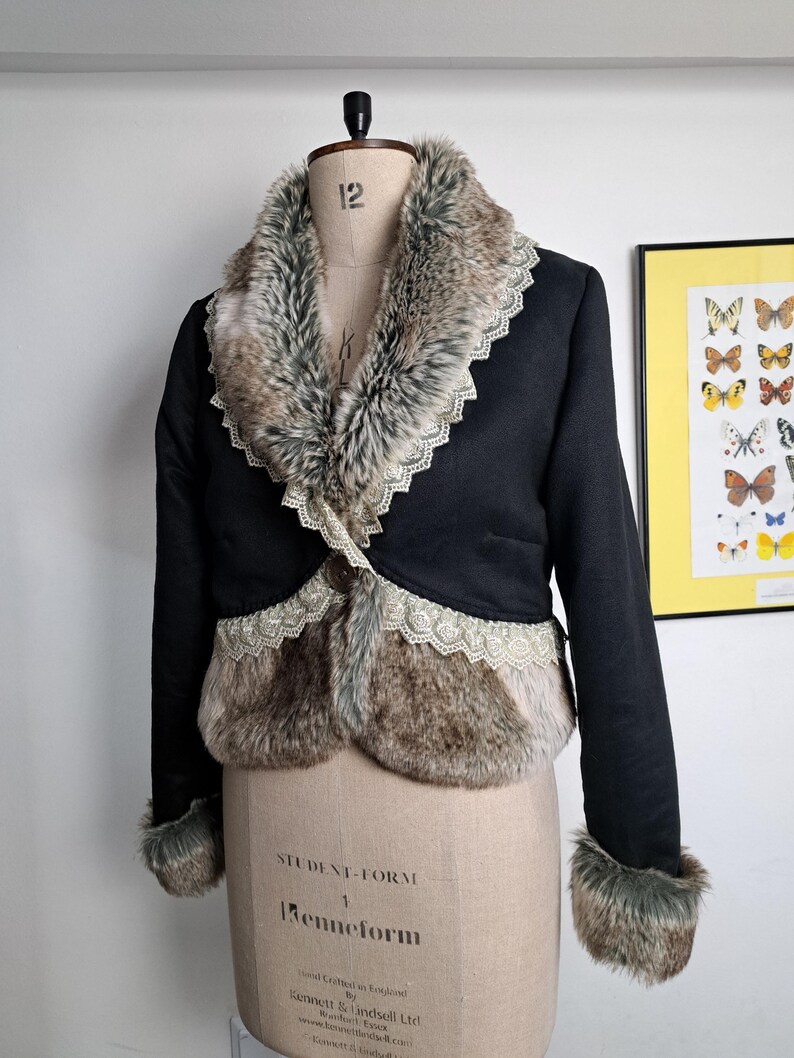 K&ouml;nnte beinhalten: Schwarze Jacke mit Kunstfellbesatz und Kragen. Die Jacke hat eine dekorative Spitzenborte entlang der Kanten des Fells und einen einzelnen Knopfverschluss. Die Manschetten sind ebenfalls mit Kunstfell besetzt. Die Jacke wird auf einer Schneiderpuppe pr&auml;sentiert.