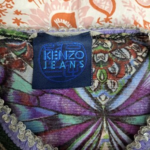 May include: A Kenzo Jeans garment featuring a dark blue rectangular label with the text KENZO JEANS in blue. The fabric displays a vibrant, multi-colored pattern with purple, green, and orange tones, finished with a light purple sequined trim.