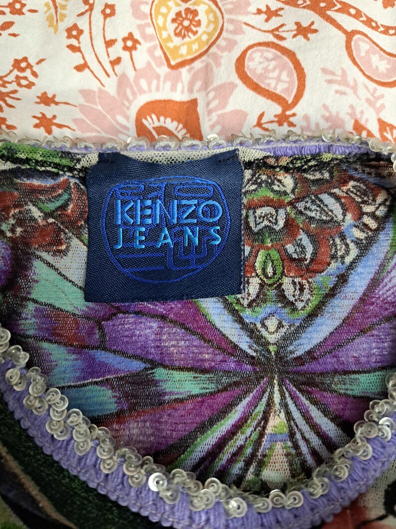 May include: A Kenzo Jeans garment featuring a dark blue rectangular label with the text KENZO JEANS in blue. The fabric displays a vibrant, multi-colored pattern with purple, green, and orange tones, finished with a light purple sequined trim.