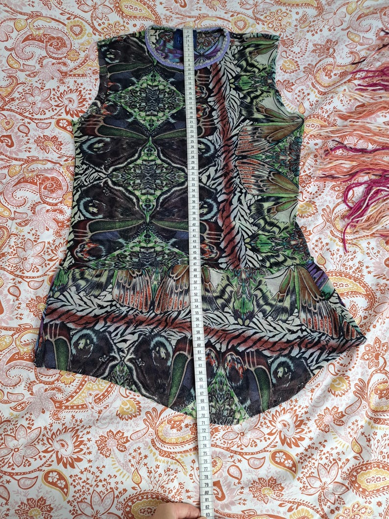 May include: A sleeveless top featuring a repeating butterfly wing pattern in shades of green, black, and brown, with a light purple trim at the neckline. A white measuring tape is placed vertically over the center, showing a length of 75 centimeters.