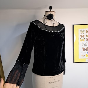 May include: Black velvet top with long sleeves and a sheer mesh neckline. The sleeves feature a sheer lace cuff with button details. A black rose choker is around the mannequin's neck. The top is displayed on a mannequin.