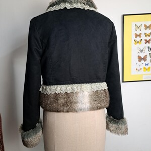K&ouml;nnte beinhalten: Schwarze, kurze Jacke mit Kunstfellbesatz an Kragen, Manschetten und Saum. Die Jacke hat eine Spitzenborte am Saum und Kragen. Die Jacke wird auf einer Schaufensterpuppe pr&auml;sentiert.