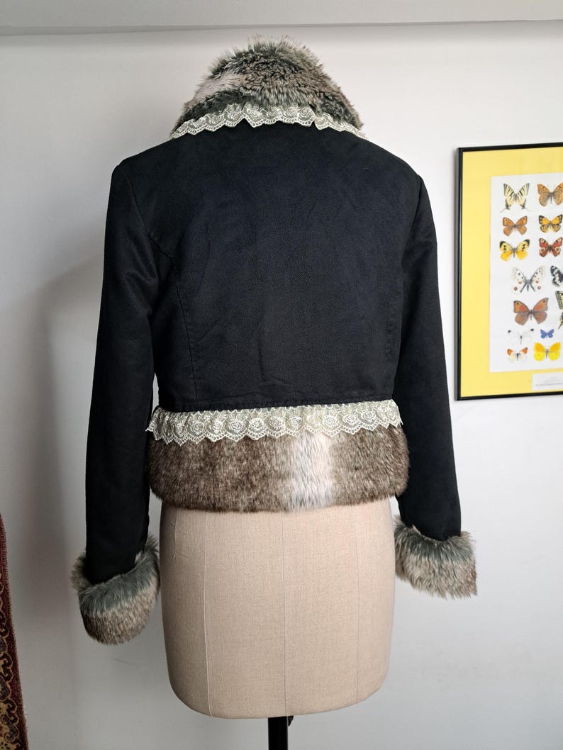 K&ouml;nnte beinhalten: Schwarze, kurze Jacke mit Kunstfellbesatz an Kragen, Manschetten und Saum. Die Jacke hat eine Spitzenborte am Saum und Kragen. Die Jacke wird auf einer Schaufensterpuppe pr&auml;sentiert.