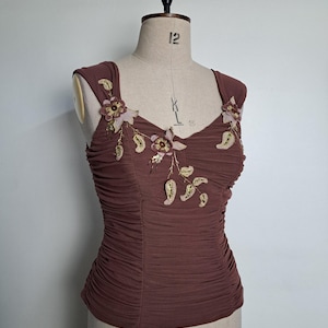 Könnte beinhalten: Ein braunes, gerafftes Oberteil mit floralen Verzierungen. Das ärmellose Oberteil hat einen herzförmigen Ausschnitt und wird auf einer Schaufensterpuppe präsentiert. Die floralen Details umfassen rosa und goldfarbene Perlenakzente, die dem Kleidungsstück einen Hauch von Eleganz verleihen.
