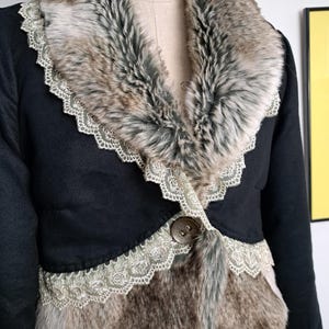 K&ouml;nnte beinhalten: Schwarze Jacke mit Kunstfellkragen und -besatz. Der Kragen und die Vorderteile weisen eine Mischung aus braunem, grauem und wei&szlig;em Kunstfell auf. Die Jacke ist mit einer zarten Spitzenborte und einem gro&szlig;en Knopf versehen.