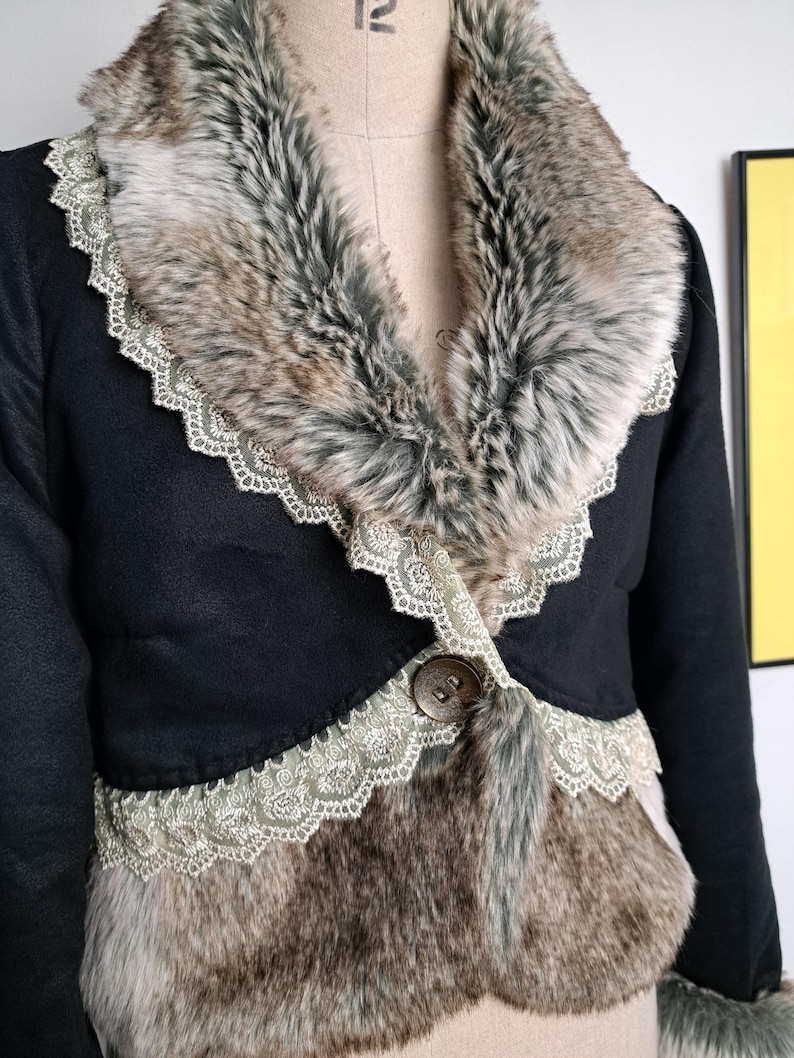 K&ouml;nnte beinhalten: Schwarze Jacke mit Kunstfellkragen und -besatz. Der Kragen und die Vorderteile weisen eine Mischung aus braunem, grauem und wei&szlig;em Kunstfell auf. Die Jacke ist mit einer zarten Spitzenborte und einem gro&szlig;en Knopf versehen.