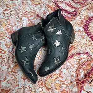 以下が含まれることがあります： シルバーの星の装飾が施された黒いアンクルブーツ。ブーツはローヒールで、使い古された革のような外観です。オレンジ、ピンク、白の色合いの模様入りの生地の上に置かれています。ファッションアイテムです。