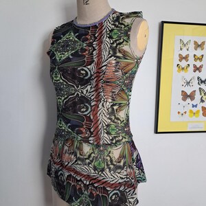 May include: A sleeveless top featuring a vibrant, multi-colored butterfly wing pattern in shades of green, orange, black, and white. The garment has a round neckline with a light purple trim and a peplum-style hem, displayed on a dress form.