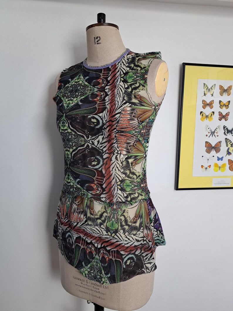 May include: A sleeveless top featuring a vibrant, multi-colored butterfly wing pattern in shades of green, orange, black, and white. The garment has a round neckline with a light purple trim and a peplum-style hem, displayed on a dress form.