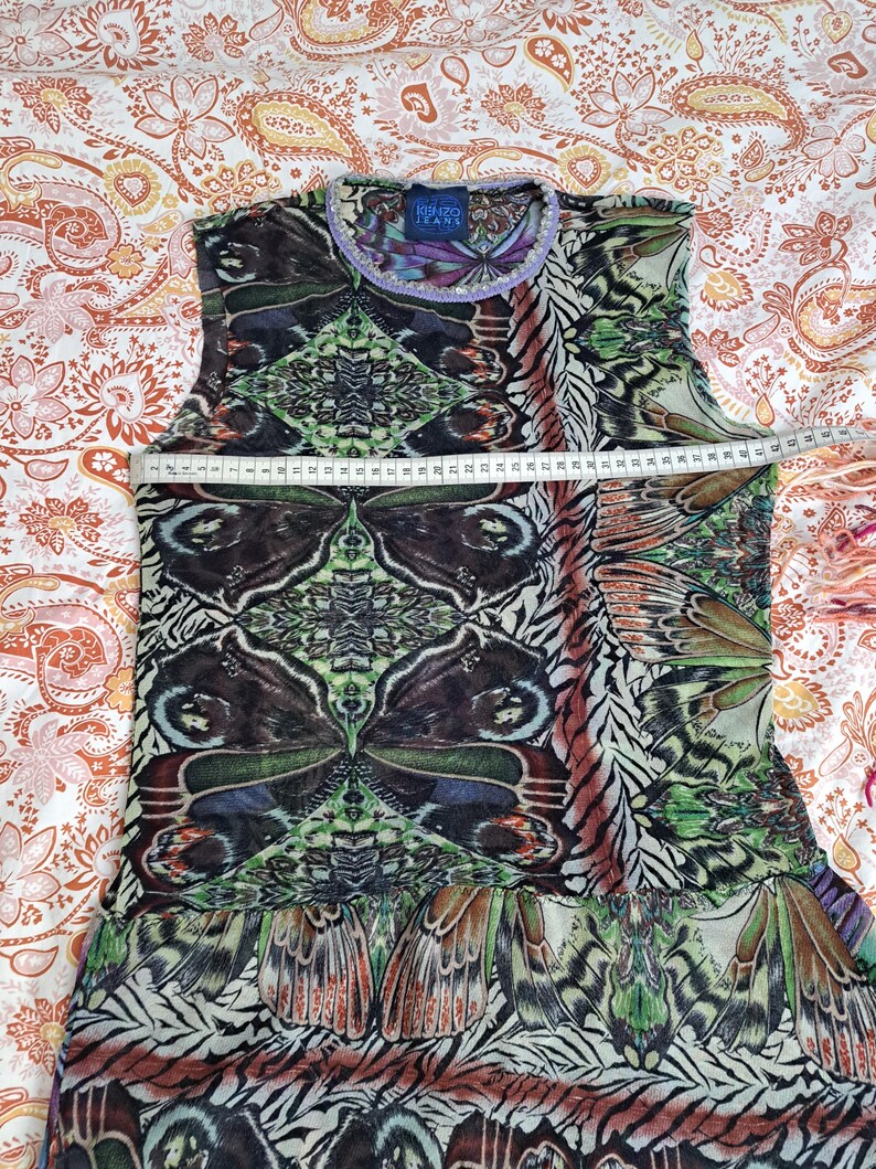 May include: A sleeveless top featuring a vibrant butterfly wing print in shades of green, brown, and black. A white measuring tape is placed across the chest, showing a width of approximately 41 centimeters. The garment has a round neckline with a light purple trim and a Kenzo Jeans label at the collar.