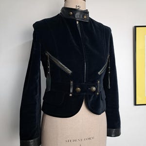 Può includere: Giacca in velluto nero con colletto alto e dettagli in pelle. La giacca presenta una chiusura con cerniera, cerniere decorative e dettagli con bottoni. La giacca è esposta su un manichino.