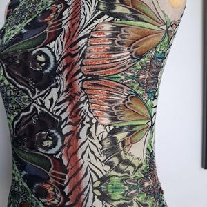 May include: A sleeveless top featuring a vibrant, repeating pattern of butterfly wings in shades of green, brown, orange, and black. The garment has a high neckline with a thin purple trim and a fitted silhouette, showcasing intricate wing details.