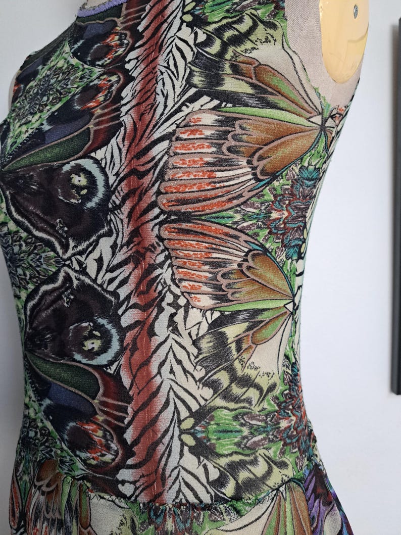 May include: A sleeveless top featuring a vibrant, repeating pattern of butterfly wings in shades of green, brown, orange, and black. The garment has a high neckline with a thin purple trim and a fitted silhouette, showcasing intricate wing details.