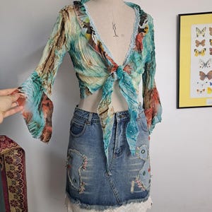 May include: A long-sleeve, tie-front crop top in a crinkled, multi-colored fabric with teal, brown, and orange patterns, paired with a blue denim mini skirt featuring decorative stitching, small colorful accents, and a white lace trim hem.
